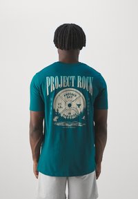 Tyrkysové bavlnené tričko s grafickým dizajnom s váhovou doskou a prírodnou scénou, s textom "PROJECT ROCK" a "ANOTHER DAY IN PARADISE".