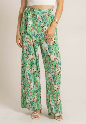 Femme portant un pantalon à jambes larges avec un motif floral vert, bleu sarcelle et marron, des sandales blanches à lanières et un bracelet en or au poignet gauche.