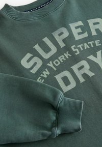 Zelen pulover s rebrastimi manšeti in izrezom, ki ima velik izbledel napis "SUPER DRY" v krem barvi, spodaj pa v manjšem fontu "NEW YORK STATE".
