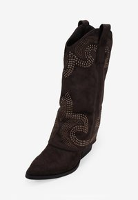Bota de vaquero de ante marrón con punta afilada, adornos metálicos decorativos que forman un patrón en espiral y un tacón bajo. Diseño clásico del cañón con lengüetas de agarre.