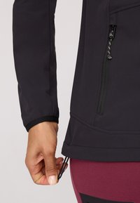 Black softshell jacket with a fitted design, elastic cuffs, side zipper pocket, and adjustable hem. Close-up of hand gripping the hem.