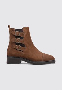 Bota de ante marrón de tobillo con tres hebillas decorativas, paneles laterales elásticos, puntera afilada y tacón bajo apilado. Textura suave y líneas limpias.