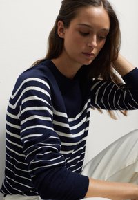 Maglione a righe blu navy e bianco in maglia con scollatura rotonda e polsini a costine. Texture morbida, design aderente. Indossato con pantaloni chiari.