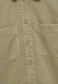 Gros plan sur le devant d’une chemise beige à boutons montrant quatre boutons et deux poches poitrine avec des coutures apparentes sur un tissu texturé.