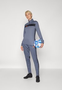 Chándal deportivo gris con parte superior de media cremallera, panel de acento negro y balón de fútbol azul. Presenta detalles de tres rayas negras y zapatillas negras.