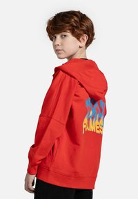 Czerwona bluza z kapturem w luźnym kroju, z dużą grafiką płomienia na plecach w niebieskim i żółtym kolorze. Miękki materiał i przestronny kaptur.