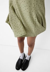 Abito texturizzato verde chiaro con piccoli punti neri, abbinato a mocassini neri e calzini bianchi alla caviglia. Visibile all'altezza della coscia.