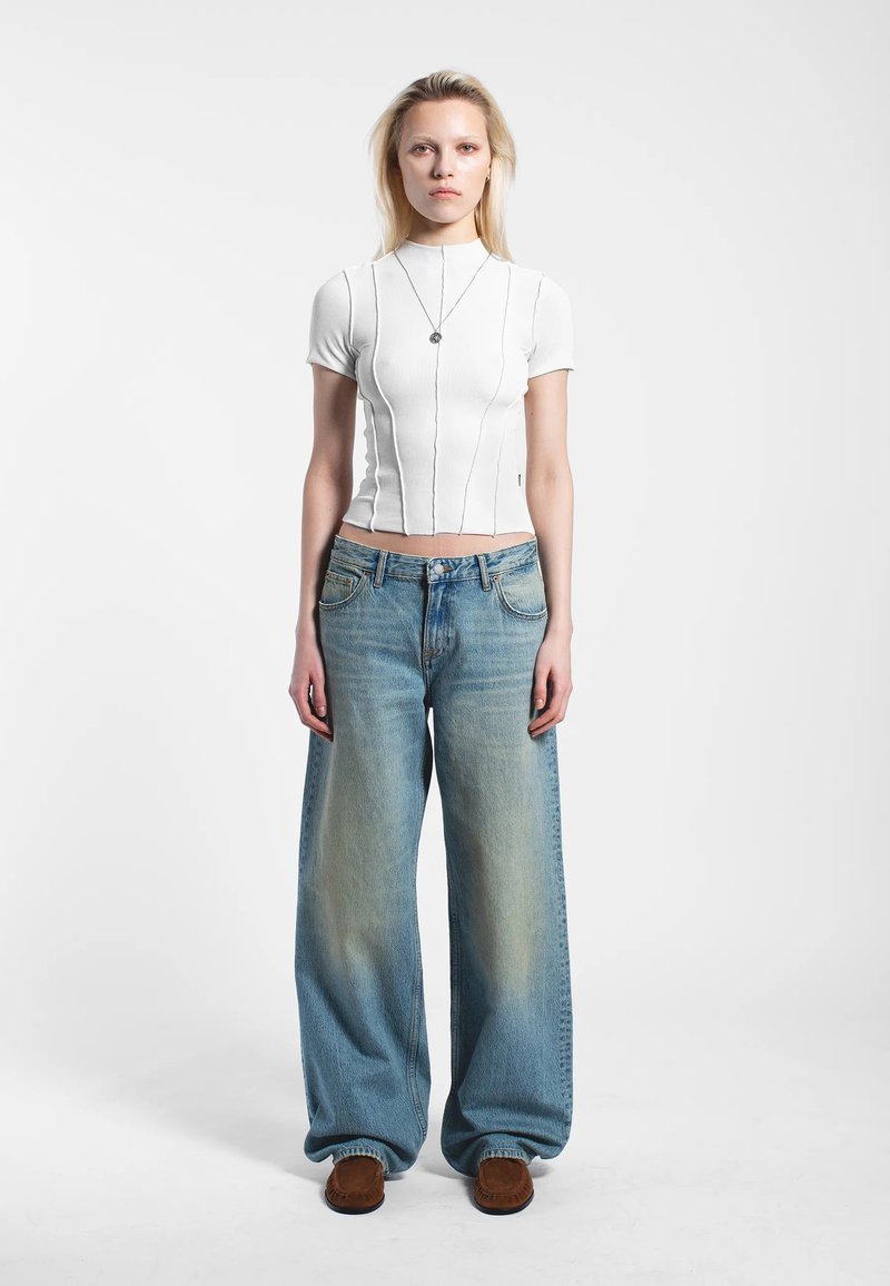 Vit ribbad topp med korta ärmar och vertikala sömmar; ljusblå vida jeans med blekta områden; bruna slip-in-skor. Enkel silverhalsbandsdetalj.