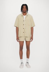Helles beige Kurzarmhemd mit braunen Knöpfen und einer Brusttasche, kombiniert mit passenden Shorts. Dazu werden weiße Socken und Sneaker getragen.
