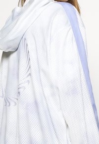 Síťovaná bunda v bílé barvě s jemnými fialovými akcenty. Obsahuje kapuci, prodyšnou texturu a decentní abstraktní vzor na zádech.