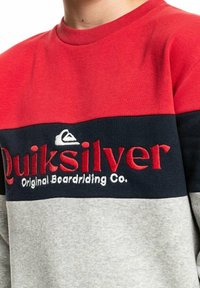 Sudadera roja, azul marino y gris con cuello redondo. Lleva un logo y la marca "Quiksilver" bordados. Material suave de mezcla de algodón, corte casual.