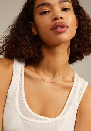 Goldene Halskette an einer Frau mit lockigem Haar, die ein weißes Tanktop mit einem weichen, strukturierten Stoff und einem mehrlagigen Halsauschnitt trägt.