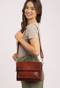 Giovane donna sorridente, con un top beige e pantaloni verdi, che porta una borsa a tracolla in pelle marrone con motivo coccodrillo.