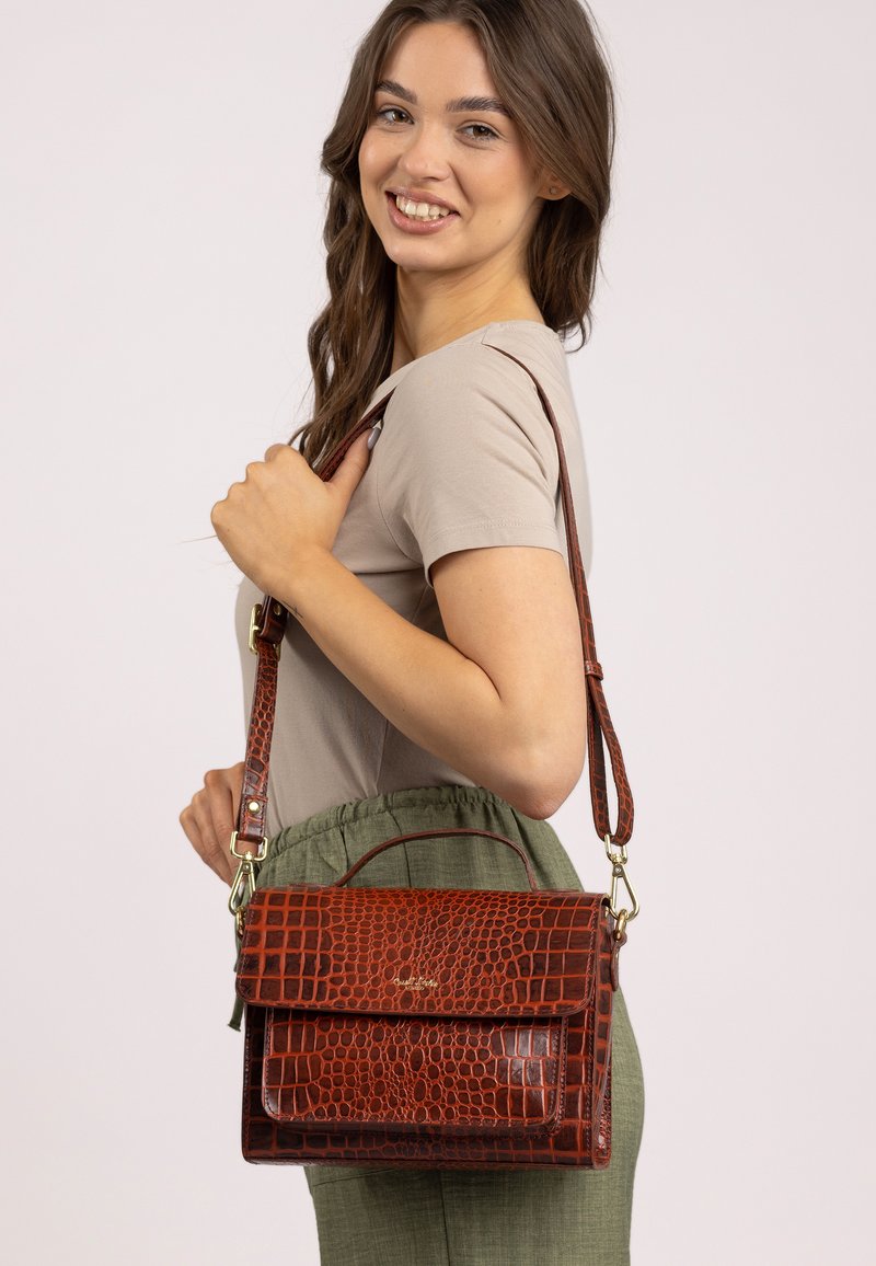 Giovane donna sorridente, con un top beige e pantaloni verdi, che porta una borsa a tracolla in pelle marrone con motivo coccodrillo.