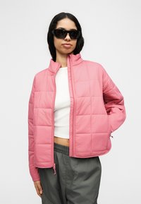 Γυναίκα με μακριά σκουρόχρωμα μαλλιά που φοράει μαύρα γυαλιά ηλίου, ροζ μπουφάν με ραφές, λευκό κοντό τοπ και σκούρο γκρι παντελόνι σε λευκό φόντο.