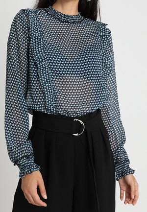 Woman wearing a long-sleeve, patterned, sheer blouse with ruffles paired with high-waisted black trousers and a belt with a circular buckle.