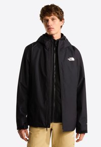 Ung mand iført en sort hættetrøje fra North Face over en sort lynlåstrøje og beige bukser, stående mod en lys baggrund.