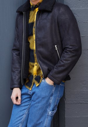 Persona che indossa una giacca nera in shearling sopra una camicia a quadri gialla e nera, con jeans blu, in piedi contro una parete grigia.