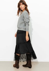 Sweat-shirt gris, jupe maxi noire superposée avec bordure en dentelle, bottes en daim marron. La jupe présente un design plissé et des détails de coutures horizontales.