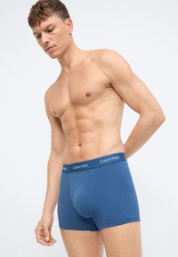 Mėlyni medvilniniai bokseriai su prigludusia forma ir Calvin Klein logotipo juosta. Audinys atrodo lygus su švelniu blizgesiu.