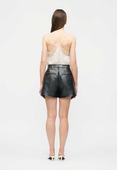 Žena s dlhými vlasmi, oblečená v bielom riasenom tielku bez rukávov, čiernych kožených šortkách a bielych topánkach na opätku, stojaca chrbtom.