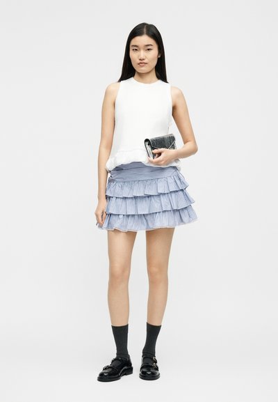 Γυναίκα που φοράει λευκό αμάνικο τοπ, μπλε φούστα με στρώσεις και βολάν, μαύρες κάλτσες, μαύρα παπούτσια, κρατώντας μαύρο τσαντάκι σε απλό φόντο.