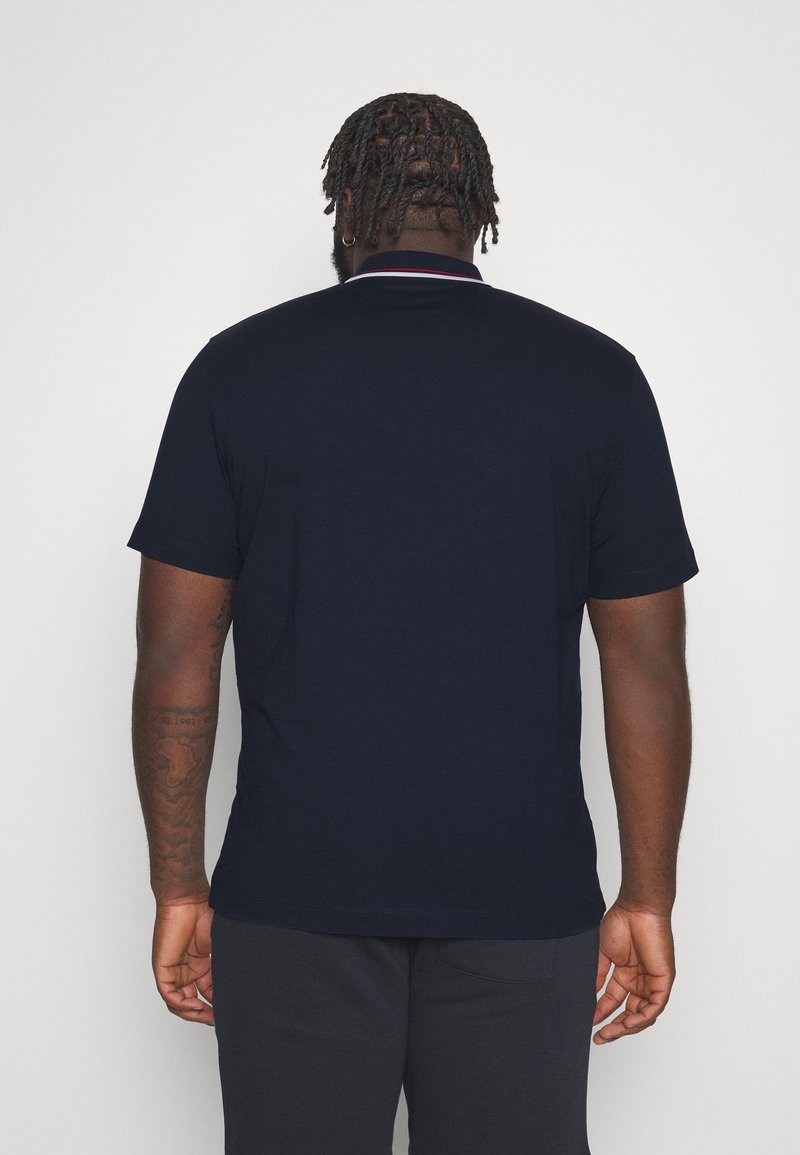 Marinblå kortärmad polotröja med en kontrasterande krage och en slät textur. Synliga tatueringar på armen, kombinerat med mörka byxor.