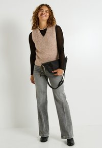 Un gilet en maille rose clair avec un col en V, associé à des manches longues noires ajustées et un jean gris évasé. Sac à bandoulière noir avec une chaîne.