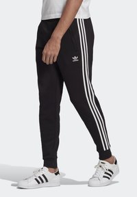 Svarta joggingsbyxor med vit tre-stripes design, med sidofickor och en broderad logotyp på vänster lår. Kombinerade med vita sneakers.