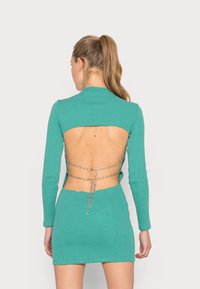 Vestido azul-petróleo com mangas longas, costas abertas e detalhe em corrente prata ligando os lados. Silhueta ajustada.