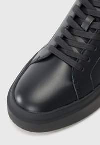 Zwarte leren sneaker met een gladde oppervlakte, ronde neus en een lage rubberen zool. Beschikt over zwarte veters en minimale stikseldetails.