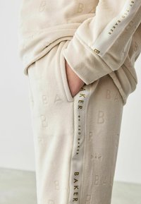 Beige fleece loungewear sæt med præget "B" mønster, sidelomme og kontrasterende hvid panel med "Ted Baker" branding i guld.