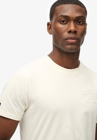 Camiseta de algodón color crema con escote redondo, mangas cortas y detalle de logo en relieve en el pecho. Textura suave y forma ajustada.