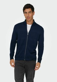 Pull à zip bleu marine avec un motif texturé, col montant et poignets côtelés, associé à un jean gris. Tissu lisse de poids moyen.