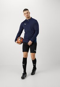 Marineblå zip-up sportjakke med hvidt logo, sorte shorts, sorte benskinner og sorte fodboldstøvler, holder en orange og sølvfaret fodbold.