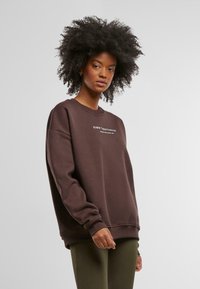 Hnědá oversized mikina s žebrovanými manžetami a lemem; na přední straně se nachází bílý textový logo. Materiál vypadá měkký a texturovaný.