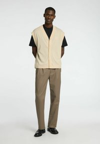 Bežna pletena brezrokavnik z V-izrezom in gumbi, oblečen čez črno srajco, v kombinaciji s khaki hlačami in črnimi čevlji. Minimalističen dizajn in čiste linije.