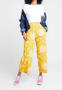 Gula blommönstrade byxor med avslappnad passform, tillsammans med en vit t-shirt och en denimjacka, avslutat med lila sneakers.