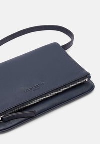 Granatowa skórzana torebka crossover o gładkiej powierzchni, z zamykaną na zamek kieszenią i wytłoczonym logo. Posiada smukły design i regulowany pasek.