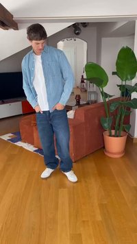Camicia a righe blu e bianche con bottoni sopra una t-shirt bianca, abbinata a jeans blu e sneakers bianche. Ambientazione interna con piante.