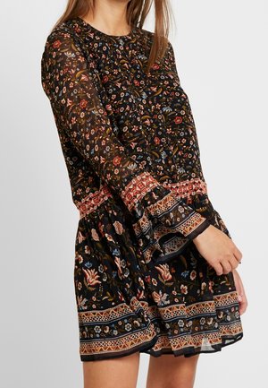 Kobieta ubrana w czarną kwiecistą sukienkę z długimi przezroczystymi rękawami oraz wzorzystymi pomarańczowo-beżowymi obramowaniami na mankietach, w talii i na dole sukienki.