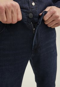 Händerna drar upp dragkedjan på mörkblå denimjeans som bärs med en mörk skjorta.
