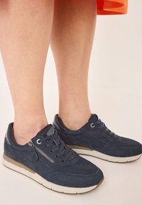 Sneakers scamosciati blu scuro con suola bianca, caratterizzati da un dettaglio con zip, design con lacci e finiture lucide sul tallone e sulla linguetta.