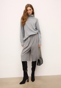 Femeie cu păr lung și ondulat, purtând un pulover gri cu guler înalt, pantaloni scurți gri până la genunchi, cizme negre cu toc înalt până la genunchi, ținând o geantă neagră cu model matlasat.