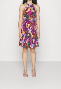 Woman wearing sleeveless purple halter dress with multicolor floral print and pink strappy sandals, standing against a plain light background.
