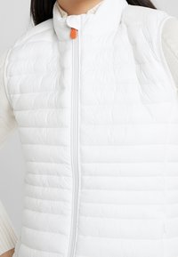 White puffer vest with a high collar, vertical quilted pattern, and a front zipper. Features an orange zipper pull for contrast.
