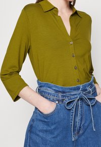 Olivengrøn skjorte med knapper og krave samt trekvartærmer, parret med høj talje blå denimbukser med et bundet bælte.