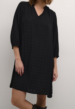 Robe noire longueur genoux avec un décolleté en V et des manches trois-quarts. Présente un motif à carreaux texturé et des fentes latérales pour faciliter les mouvements.