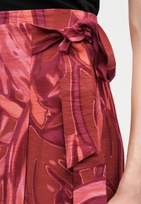 Κοντινό πλάνο στη μέση που φοράει φούστα με κόκκινο και ροζ αφηρημένο σχέδιο, με δεμένη ζώνη ύφασμα στο πλάι και μαύρο τοπ.