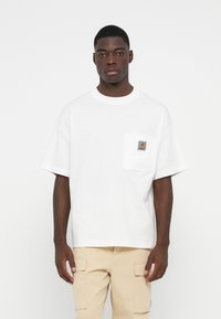 White cotton oversized t-shirt with a round neckline, short sleeves, and a chest pocket featuring a logo patch. Smooth texture, casual design.